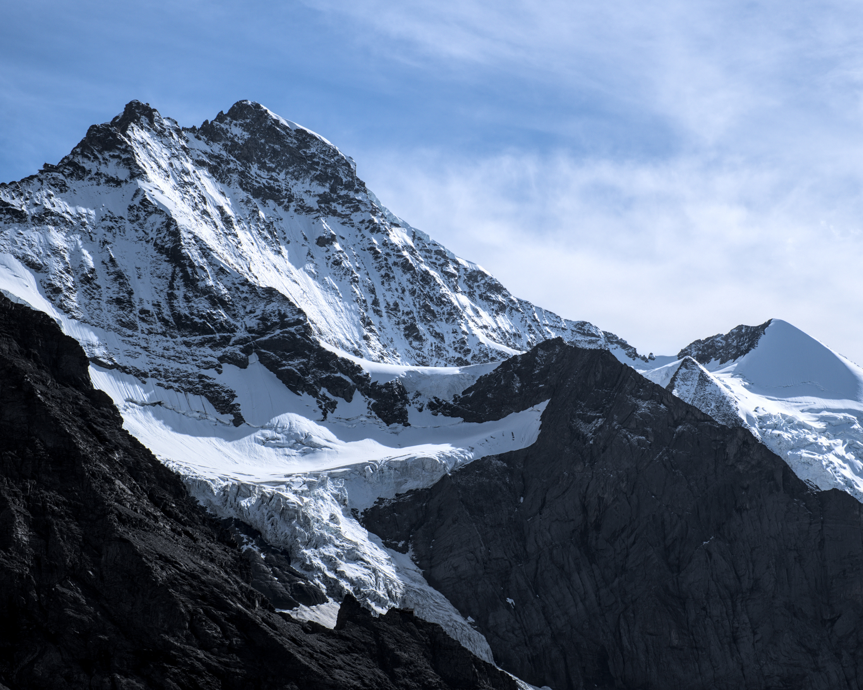A large snow covered mountain dominates the frame, with some wispy clouds in an otherwise blue sky. The rock faces are complexly textures and contain wavy bands of subtle differences in colour from what I believe is their sedimentary origins. There is a very small building visible on the ridge line at the top left of the frame, which just amplifies the sense of scale of the natural features. The scene is brightly lit, making the snow extremely bright and providing stark contrast against the almost black rock. There is another slope visible in the bottom left foreground that forms one edge of the glacier in the center of the frame, where we can see the dirtier ice of the terminus.