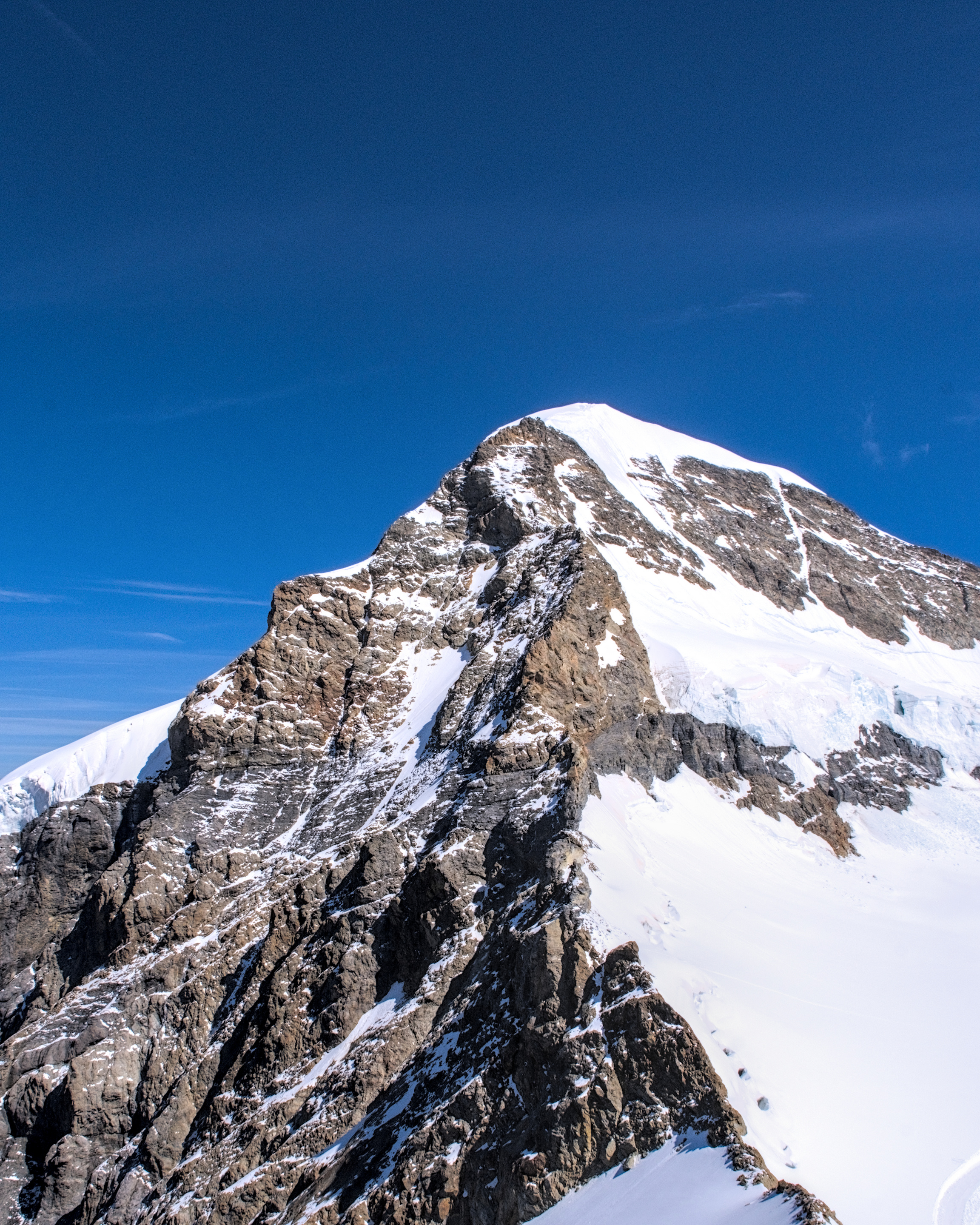 This largely brown rocky mountain occupies the bottom two thirds of the frame, with it's summit capped in snow. It's left half is mostly wonderfully textured light brown rock, with a horizontal band of striated darker grey running through roughly the middle of the visible portion. It's right half is mostly covered in brilliant white snow, showing signs of parts having sheared off in small avalanches at some point prior. There are very few shadows, but those that are visible are quite high contrast, as this face of Mönch was being directly light by the bright August midday sun. The sky is a grainy blue that gradually darkens towards the top of the frame, with barely a few wisps of cloud visible.