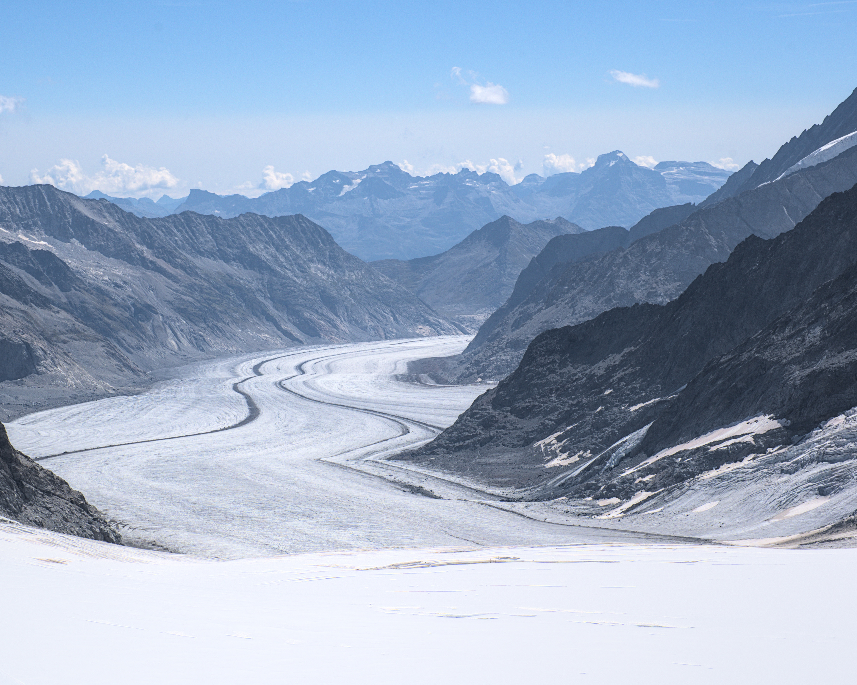 A bright white, subtly crevassed snow covered glacier occupies the bottom 1/5th of the frame. Beyond it stretches an enormous valley formed of rocky, dark grey and light brown mountains bordering the ribbon of crusty white and grey glacier ice that stretches off into the distance. A wavy darker line runs almost parallel to the border of the ribbon on either side, seeming to grow closer together as the perspective shifts with distance. The two valley sides and a pyramidal mountain converge on a point just off center of the frame where the glacier then disappears from view behind the mountains to the right. The pyramidal shaped mountain at the end of the visible section of glacier has a barely visible small building near it's summit. The sky is a pale blue, with two fluffy clouds sat above a backdrop of a hazy mountain range that almost forms a smiley face with the shape of the ridge line. There are a scattering of lower level fluffy white clouds closer in altitude to the summits of these mountains in the far distance, but none are occluding the summits.